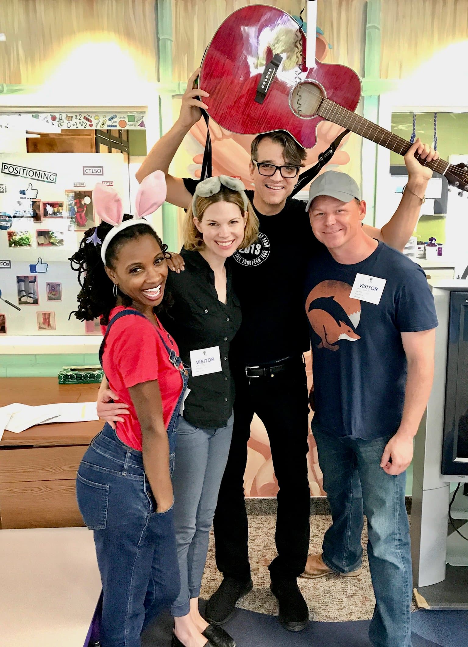 Happy Montana, in glasses and a black T-shirt, holds a red acoustic guitar over his head, smiling beside three fellow volunteers wearing visitor badges in a brightly painted room.